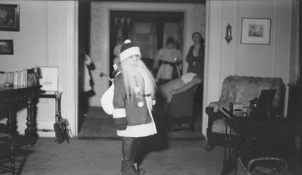 A child dressed as Santa Claus is standing and posing in the living room of the Herbert P. Brumder residence at 2030 East Lafayette Place. The costume includes boots, a false beard and a pack of toys. There are women standing in a room seen through the doorway in the background. Written on the negative envelope is: "Dress-up Skating Party."