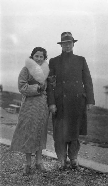 Herbert Paul Brumder and his wife, Margaret Bouer Brumder, posing outdoors standing on a hill overlooking a body of water. Both of them are wearing long winter coats, hats and gloves. There is a rustic bench in the background on the left.