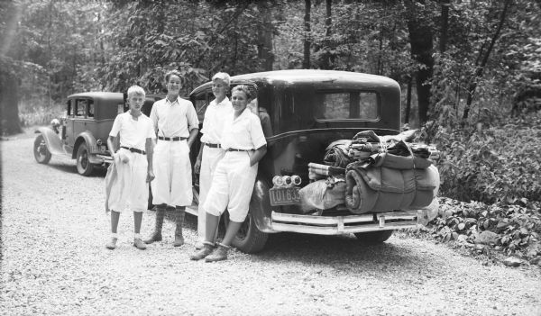 Four boys wearing light-colored summer clothes are posing beside a car with camping gear tied above the rear bumper. There is a second car parked ahead of the loaded car. Three of the boys are wearing knickerbockers. The boys include Philip Brumder, far left, and his brother Herbert E. Brumder, second from right, wearing long pants. The two other boys, possibly cousins, are unidentified. They are standing on a gravel road or drive in a wooded area.