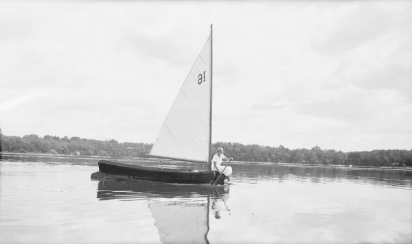 Barbara Brumder, wearing a light-colored dress, is sitting on the bow of a dinghy sailboat, paddling across the glassy waters of Pine Lake. She is the daughter of Herbert P. and Margaret Bouer Brumder.