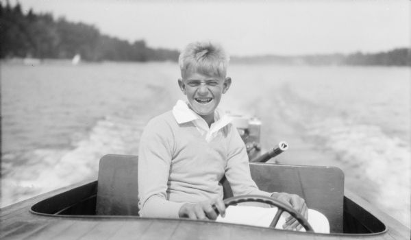 Herbert E. Brumder squinting and smiling broadly while sitting behind the wheel of a Chris-Craft wooden power boat on Pine Lake. He is wearing a light sweater and shirt. In the background is the boat's wake, and the shoreline.