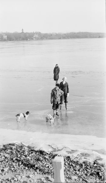An elevated view from the shore of frozen Pine Lake featuring Philip Brumder, front, and his sister Barbara, with their mother Margaret in the rear. All are wearing ice skates. There are two dogs on the ice in the foreground. Two large houses and an observation tower are visible on the far shoreline.