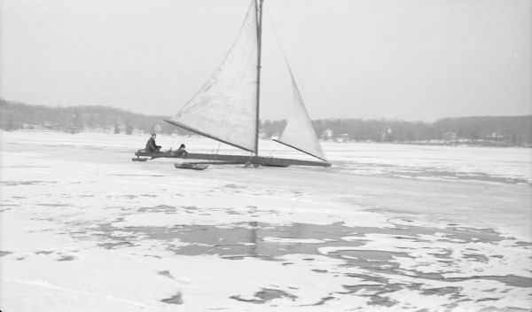 Two unidentified men sail their ice boat across the frozen surface of Pine Lake.  A light snow covers much of the ice.