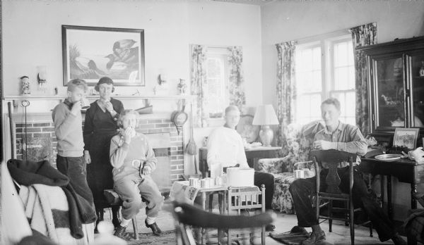 A family group poses in the living room of the Herbert P. Brumder home at Pine Lake.  From left are Philip, his mother Margaret, Barbara, and Herbert E. Brumder. The young man at far right is not identified. The group is enjoying food and drink from a small table holding tin cups, a bottle of clear liquid, and a tin container. Sweaters and coats are piled on a chair at left. A print of J.J. Audubon's <i>Canvas Back Ducks</i> is hanging over the fireplace.  