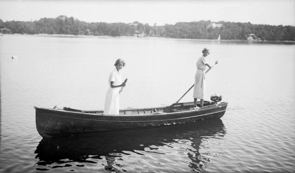 Two unidentified young women are standing in a canoe with a small motor, and each of them is holding an oar. They are wearing light-colored summer dresses. Boathouses and a large house are on the far shore.