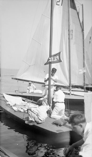 Three young men are working on a racing sailboat which is flying a P.L.Y.C. (Pine Lake Yachting Club) pennant. Other boats are moored alongside.