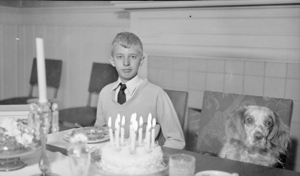 Philip George Brumder, son of Herbert P. and Margaret Bouer Brumder, sitting at the dining table in the family home at 2030 East Lafayette Place. He is wearing a pullover sweater, shirt and tie. A dog is sitting on the chair beside him. On the table, candles are burning on a birthday cake.