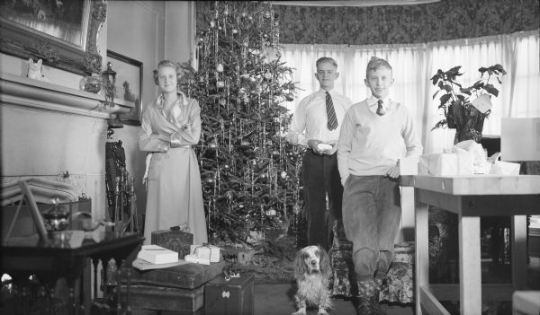 The three children of Herbert P. and Margaret Bouer Brumder, from left, Barbara, Herbert E. and Philip, posing standing in the living room of the family home at 2030 East Lafayette Place on Christmas morning. The family dog is standing next to Philip. Barbara is wearing a housecoat with satin lapels and cuffs. The boys are wearing neckties. A tall Christmas tree with glass ornaments and strands of tinsel is in the background, and a Poinsettia plant is at right. There are wrapped gifts on a sturdy work table at right. Near Barbara are a suitcase and a radio, as well as gifts on an ottoman.  