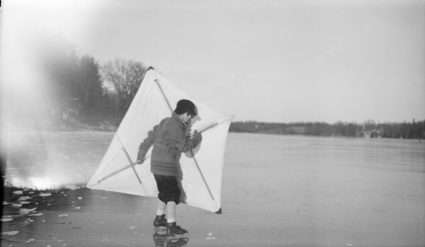 Philip George Brumder, wearing knickers, a long belted jacket and ice skates, is holding a large kite while skating on Pine Lake.