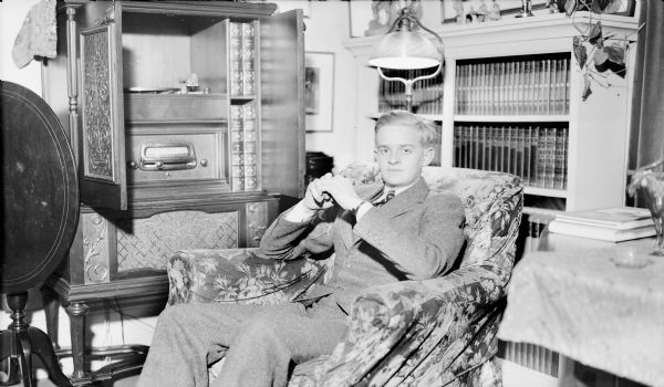 Herbert E. Brumder, oldest child of Herbert P. and Margaret Bouer Brumder, is wearing a suit and tie, and is sitting in an easy chair in front of the open cabinet of a combination radio and record player. Behind him is a floor lamp and a full bookcase.