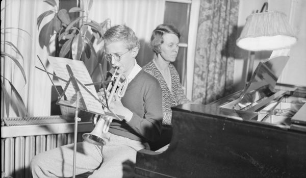 Philip George Brumder, sitting on the end of a piano bench, reads from sheet music on a stand and plays a cornet while his mother, Margaret Bouer Brumder, shares the bench and accompanies him on piano.  There is a floor lamp at right.