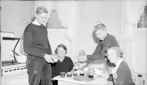Margaret Bouer (Mrs. Herbert P.) Brumder, second from left, is sitting at a small kitchen table surrounded by her children, from left, Herbert E., Philip, and Barbara. They are coloring Easter eggs. A large pan is sitting on the stove behind Herbert.