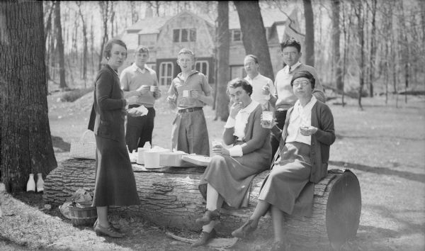 Mararet Bouer (Mrs. Herbert P.) Brumder, far left, and her son Philip, third from left, join other friends or relatives gathered around a large fallen log to enjoy a picnic. A picnic basket, marshmallow tins, and a metal box with hinged lid are resting on the log; there are two milk bottles against the tree trunk at left. Herbert P. Brumder's Pine Lake summer house is in the background.