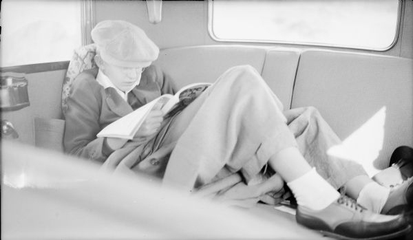 Philip George Brumder reading a magazine in the back seat of a car, while leaning against the side with his feet up on the seat. He is wearing a cap and has an overcoat across his lap and legs.