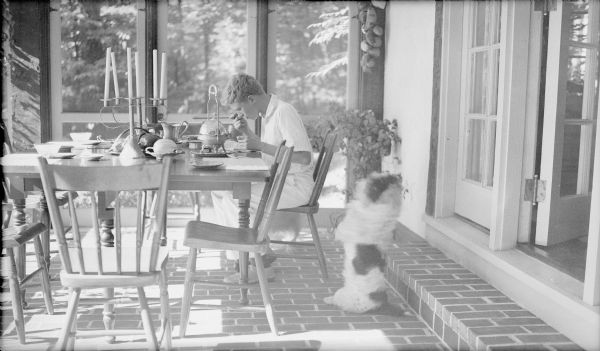 The photographer's son, Herbert E. Brumder, enjoying breakfast on the east-facing screened porch of the family's summer home on Pine Lake. The family dog "begs" near Herbert's chair. There is a tin candelabra on the table and several other places are set. The porch has a brick floor, with a step leading to an open French door into the house.