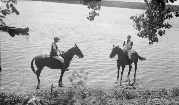 Two unidentified women riding horses with English saddles in the shallow water near the shore of Pine Lake. The women are wearing light-colored jodhpurs and tall boots. There is a canoe on the left.