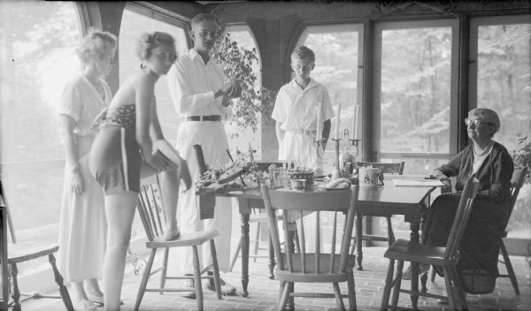 The photographer's son, Herbert E. Brumder, towers over his mother and siblings as he stands at a table which holds a birthday cake.  Pictured are, from left, Margaret Bouer (Mrs. Herbert P.) Brumder, Barbara, Herbert, and Philip Brumder, and an unidentified woman seated at the table.  The group is gathered on the screened porch at their summer home on Pine Lake.  The porch has a brick floor.