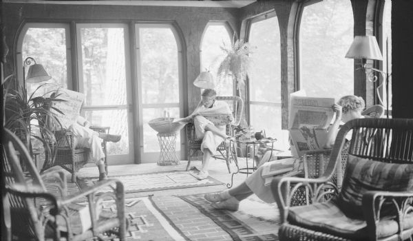 The photographer's three children, from left, Herbert E., Philip, and Barbara Brumder reading newspapers while sitting on the newly constructed screened porch of the family's summer home on Pine Lake. The boys are reading the comics from the Milwaukee Journal while Barbara is holding <i>The American Weekly</i>, a syndicated Sunday supplement published by the Hearst Corporation. The porch has unfinished rough-hewn beams, and is furnished with wicker furniture.