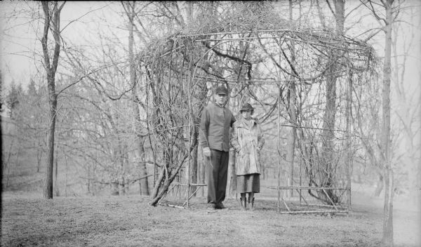 Herbert P. Brumder poses with his wife Margaret Bouer Brumder in the doorway of a metal framed gazebo. Large vines cover the framework. The gazebo was on the lawn of Villa Henrietta, the summer estate of Herbert's father, Milwaukee publisher and businessman George Brumder.