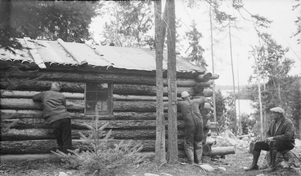 A well-dressed man sitting on a boulder at right is watching two other men repair the chinking on a log cabin. Tar paper roofing has been applied over the original log roof. There is a lake in the background. The man at far left is likely Herbert E. Brumder, the photographer's son.