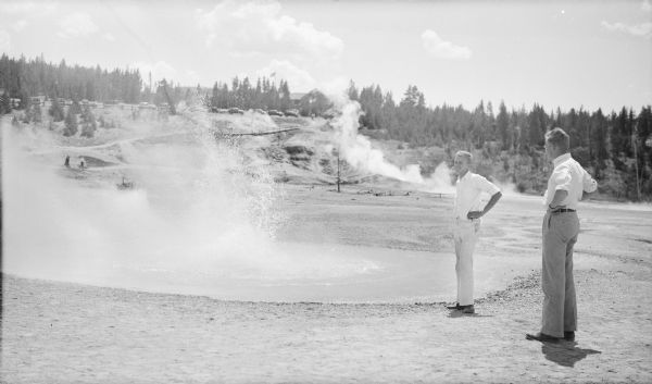 The photographer's two sons, Philip, left, and Herbert E. Brumder are standing near a steaming pool in one of Yellowstone's geyser basins. There are cars parked along a low fence in the background, near a lodge.