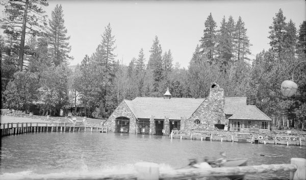 A wooden power boat is leaving a wake in the foreground of a view of a rustic stone "wet" boathouse with four stalls for boat storage. The building also has a massive chimney and a sun room at right. There is a second stone cottage or lodge in the background, left. The buildings are set among tall trees. In the right foreground is a Japanese lantern style light fixture.  