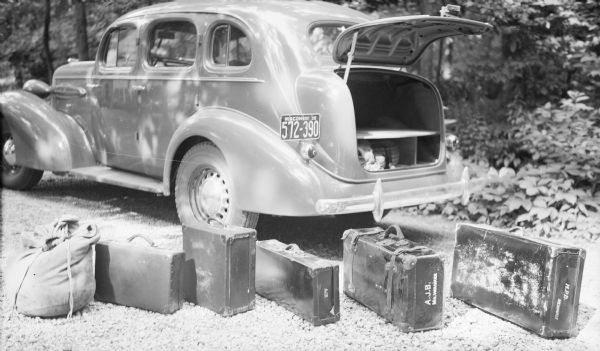 Five suitcases and a duffle bag are lined up on a gravel road near the open trunk of a 1935 Oldsmobile four door sedan. The highly polished car is parked in dappled shade in a wooded area. Two suitcases bear the monogram A.J.B., and a third is marked H.P.B. (Herbert Paul Brumder) Milwaukee.  