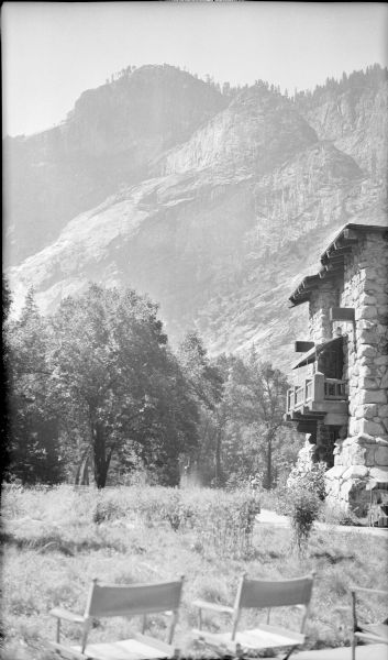 At right, the side of the Ahwahnee Hotel, a stone building, helps frame a view of granite mountains in Yosemite National Park. A balcony extends from the wall. Canvas director's chairs in the foreground face the view.