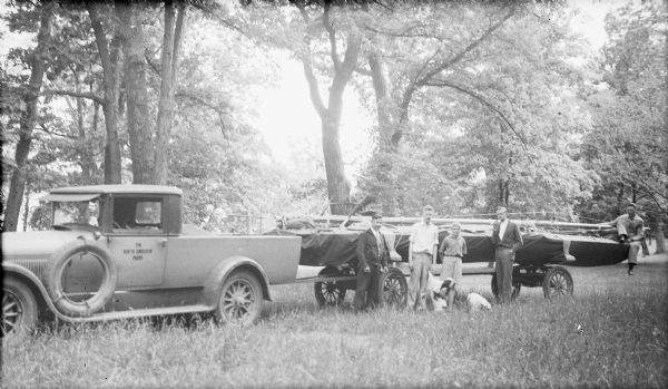 A circa 1930 pickup truck pulling a trailer with a racing yacht is parked in a grassy, wooded area. "The North American Press" is printed on the truck's door. An unidentified man in work clothes is sitting on the boat at far right. Second from right is the photographer's son, Herbert E. Brumder. Another son, Philip Brumder, is standing second from left. A dog is supporting itself against Philip's leg. There is a second dog on the ground. Two other young men are not identified.
