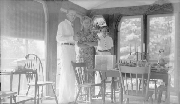 Herbert Edmund Brumder standing with his sister Barbara, center, and mother, Margaret Bouer (Mrs. Herbert P.) Brumder near the dining table on the screened porch of their Pine Lake summer home. A birthday cake with eighteen candles is on the table, which also holds a candelabra and a map.