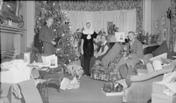 Barbara Brumder, standing center, is modeling a furry hood and is resting her left hand on her mother Margaret's head. Philip Brumder is standing at left; his brother Herbert E. Brumder, wearing a robe with plaid collar and cuffs, is sitting on the arm of a chair at right. A tall Christmas tree is standing near the bow window, and the family dog is under the tree. Boxes, wrapping paper and gifts are strewn over the furniture.