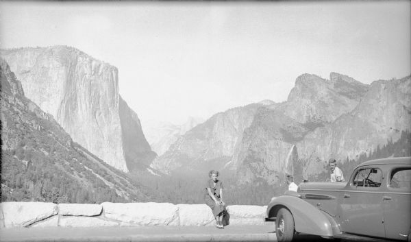 The photographer's wife, Margaret Bouer Brumder, is sitting on a low stone wall at an overlook in Yosemite National Park, with the granite formation El Capitan on the left. Son Herbert E. Brumder is sitting on the wall facing the view, while his brother Philip is facing the camera over the hood of the family car.