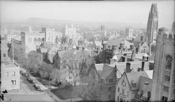 An elevated view of the Old Campus area of Yale University, with Chapel Street crossing diagonally at lower left. There is a partial view of Bingham Hall at far right. Vanderbilt Hall, with four crenelated towers and multiple chimneys, is next to it. Further along Chapel Street is Street Hall. The Yale University Art Gallery is at far left. The tallest structure in the background, right, is Harkness Tower.