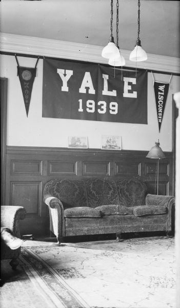 A well worn sofa and oriental-style rug furnish a high-ceilinged parlor in a dormitory at Yale University. Above the sofa is a large banner reading "YALE 1939" flanked by two pennants. The pennant at right has a seal and the letters M.C.D.S., representing Milwaukee Country Day School; at left is a "WISCONSIN" pennant. The photographer's son, Herbert E. Brumder, was a freshman at Yale University when this photograph was taken.
