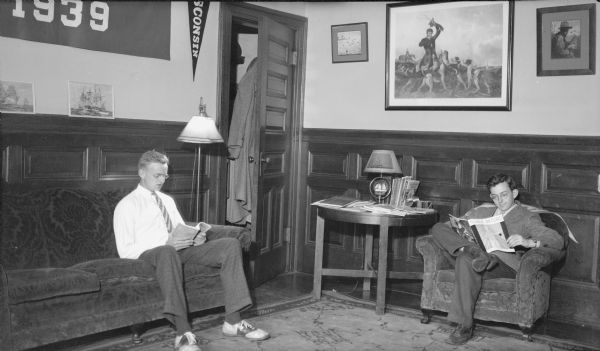Herbert E. Brumder, left, and an unidentified young man are sitting and reading in a dormitory parlor. Coats are hanging on the back of a door at center. There is a print of a hunting scene hanging on the wall.