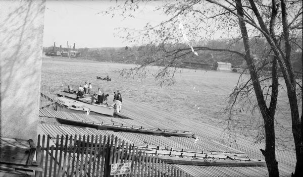 Three racing shells are resting on the ramp of the Yale boathouse on the Housatonic River. Men have gathered around two of three power boats moored at the end of the ramp. A man is rowing a canoe in the background. On the far side of the river at left is a group of industrial buildings with several smokestacks and water towers.