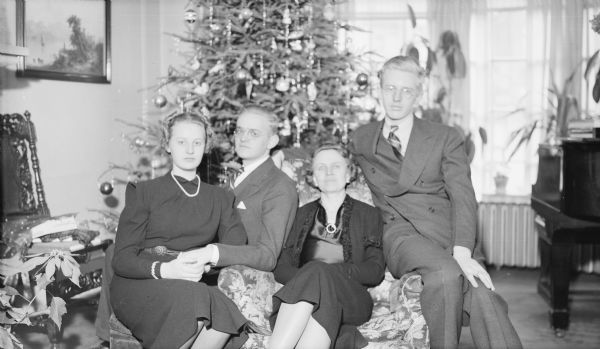 Margaret Bouer (Mrs. Herbert P.) Brumder, is sitting in an easy chair, posing with her three children in the living room of their home at 2030 East Lafayette Place. From left are: Barbara, Herbert E., and Philip.. A Christmas tree is standing behind them, and there is a poinsettia plant at far left.