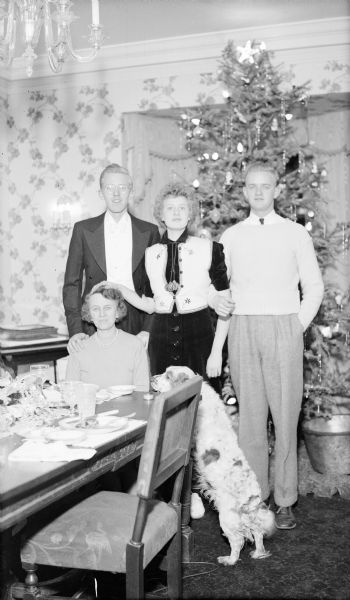 Margaret Bouer (Mrs. Herbert P.) Brumder sitting at the end of the dining table as her three children. From left are: Philip, Barbara, and Herbert E. posing standing behind her. Barbara is resting her right hand on her mother's head. Philip is wearing a tailcoat and white tie while the others are less formally dressed. The family dog is standing at the table, surveying the remains of the meal. There is a tall Christmas tree in the background.