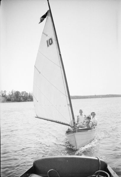 A smiling Philip Brumder, rear, poses with his thumbs tucked in his shirt in a boastful gesture as he is towed in a dinghy sailboat with his sister Barbara and an unidentified boy. The boy is holding a sailing trophy. The east shore of Pine Lake is in the background. 