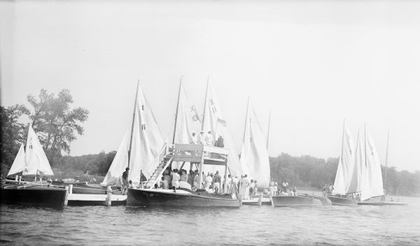 View across water towards five wooden power boats moored at a pier on Pine Lake. On the far side and at right are a number of racing yachts, with a dinghy sailboat at far left. There are four men on a raised platform on the pier, with a crowd on the pier itself.