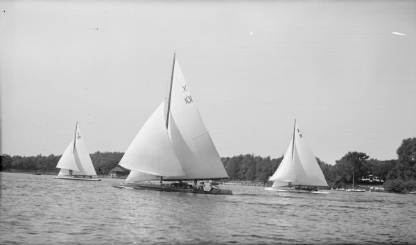 Three racing yachts flying spinnakers compete on Pine Lake. A boathouse is on the shoreline in the background, and a house and wharf are at right. 