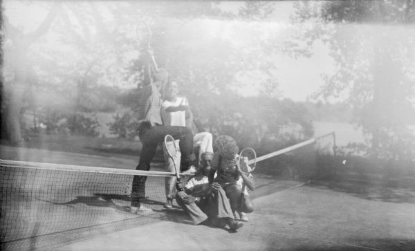 The three children of Herbert P. and Margaret Bouer Brumder are posing with an unidentified young man (rear, in white shirt) on the tennis court at their summer home. Pine Lake is in the background. Philip George Brumder stands at left with his racquet raised and his foot pushing down on the net. Herbert Edmund Brumder is sitting with his back to the net with his sister Barbara leaning against him.  