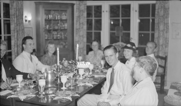 Members of the extended Brumder family have gathered in the dining room of the Herbert P. Brumder summer home at Pine Lake. The table is set for a meal and there is a birthday cake with lighted candles at the far end of the table. Philip Brumder is at far left. His brother, Herbert E. Brumder is at far right, leaning back from the table, and sister Barbara, wearing a hat, is next to him. Their mother, Margaret Bouer (Mrs. Herbert P.) Brumder is sitting at the head of the table. The woman in the foreground, right, may be Amelie Brumder Mayer. The other people are unidentified.