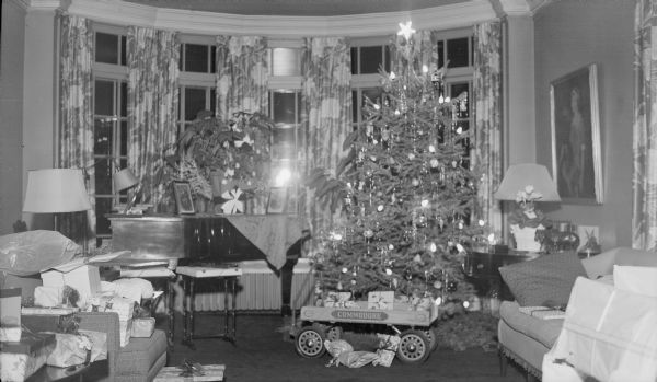 Wrapped gifts are piled on furniture and in a "Commodore" wagon in the Herbert P. Brumder residence at 2030 Lafayette Place. There is a tall  lighted Christmas tree at right, and a poinsettia plant on the piano in the background.