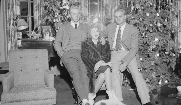 Barbara Brumder, wearing a fur coat, poses sitting in a chair flanked by her brothers, Philip, left, and Herbert E. They are the children of Herbert P. and Margaret Bouer Brumder. There is a Christmas tree at right and a poinsettia plant in the background on the left.