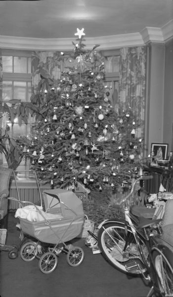 A tall, electrically lighted Christmas tree is standing in the bow window of the living room of Herbert P. and Margaret (Bouer) Brumder at 2030 East Lafayette Place. A toy baby buggy, bicycle and other gifts fill the area around the tree.