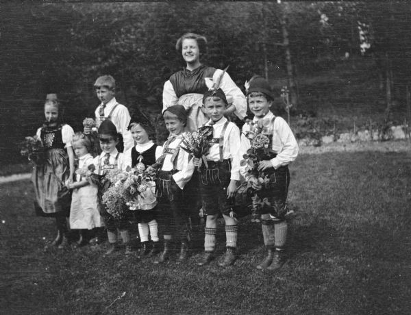 Nine children, members of the extended Brumder family, posing in traditional German costume, including lederhosen, felt caps and "Loferl" stockings for the boys, and dresses with aprons for the girls. The children are holding bouquets of spring flowers, including lilacs and lily of the valley.