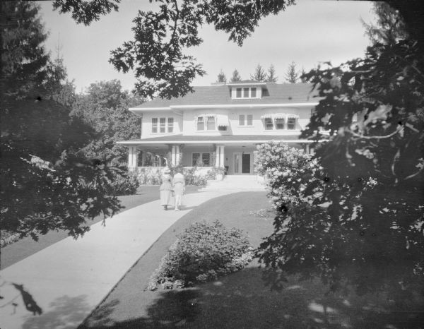Slightly elevated view of two unidentified women walking up a broad sidewalk to the lake-facing (east) facade of Villa Henrietta, originally the Pine Lake summer home of George and Henriette Brumder. The two- and one-half story house has been extensively remodeled and enlarged. There are awnings over the second story windows. A porch with double columns wraps around the front and left side of the house.