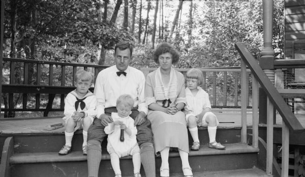 Herbert Paul Brumder, the youngest son of Milwaukee publisher and businessman George Brumder, sitting on porch steps with his wife, Margaret Bouer Brumder and their children, from left, Herbert Edmund, Philip George, and Barbara. There is a wooded area in the background.