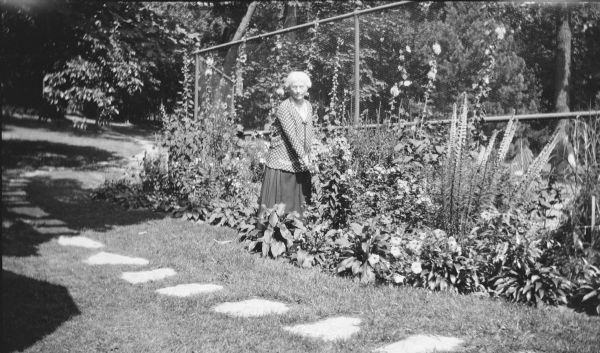 An unidentified woman posing among flowers in a border along the fenced tennis court near the summer home of Herbert P. Brumder at Pine Lake. She is wearing a skirt and polka dot jacket. There is a stepping stone path in the foreground.